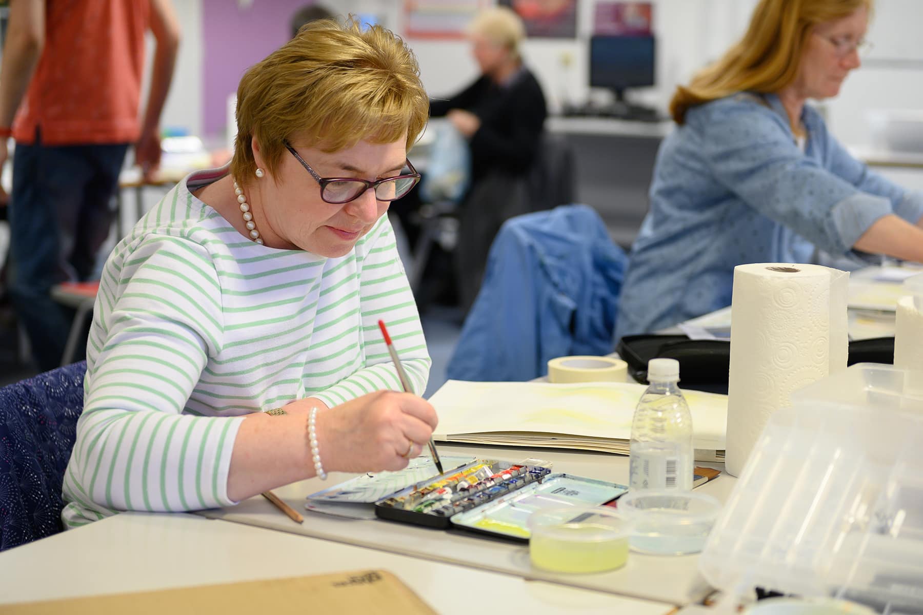 A part time leisure student sits at a table with watercolours and a paintbrush.