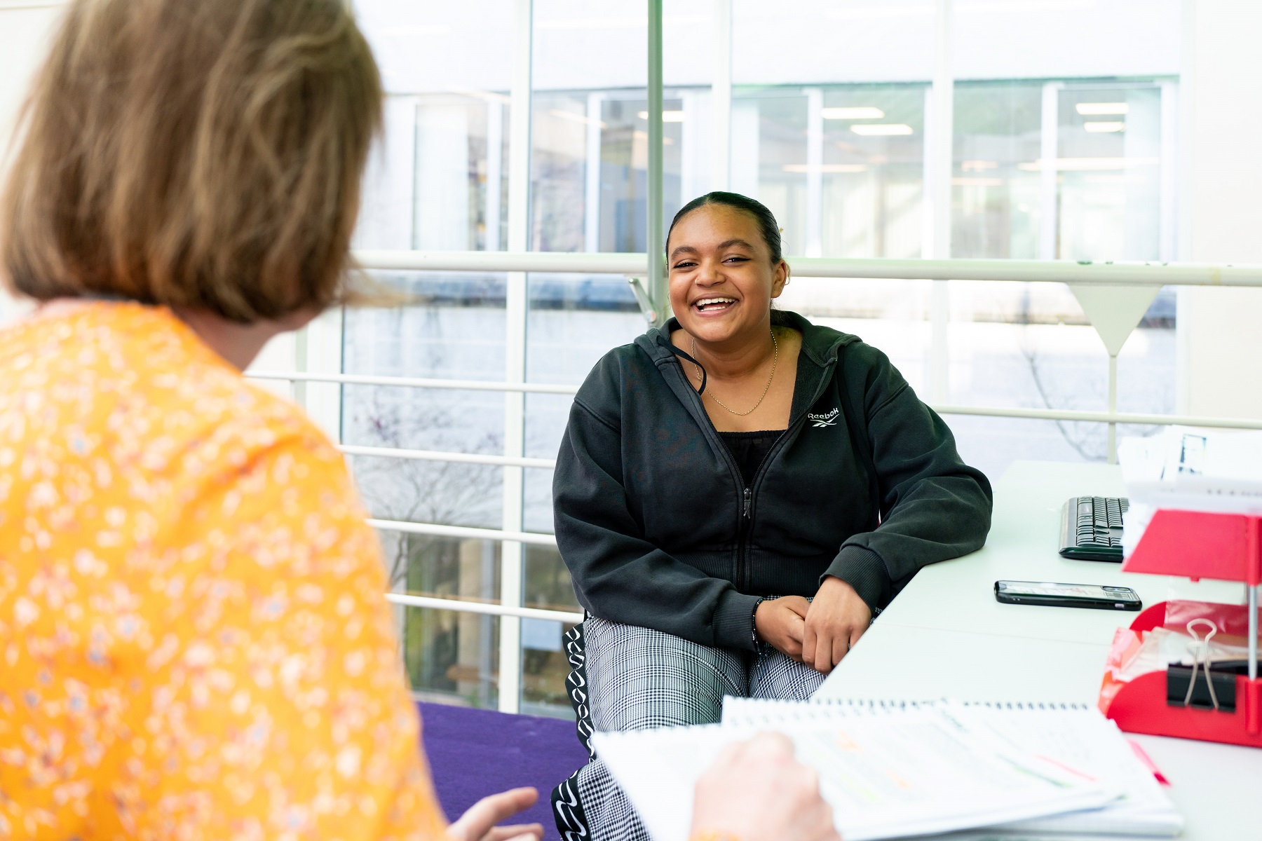 A student sits across from a member of staff. The student looks over the shoulder of the staff member towards the camera and smiles.