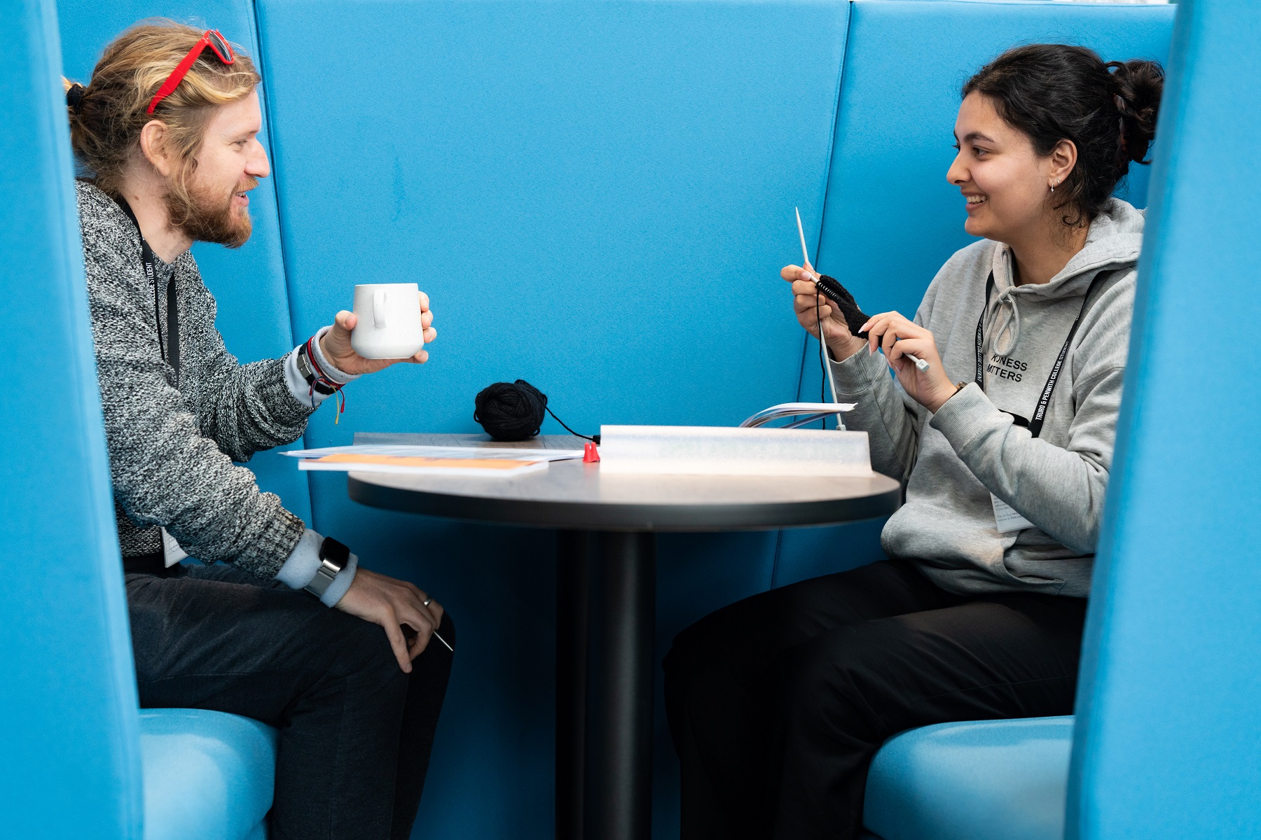 General HE 0010 Two students sit in a seating booth at the University Centre Truro and Penwith. One has a mug in their hands while the other is knitting. Both smile at each other.