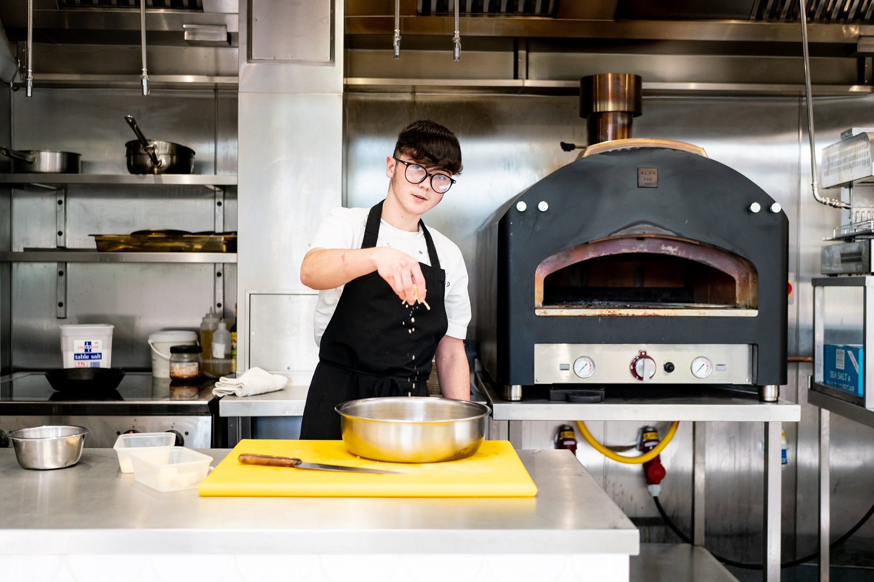 An Apprentice in a kitchen with pizza oven in the background looks down the camera while sprinkling flour into a bowl.