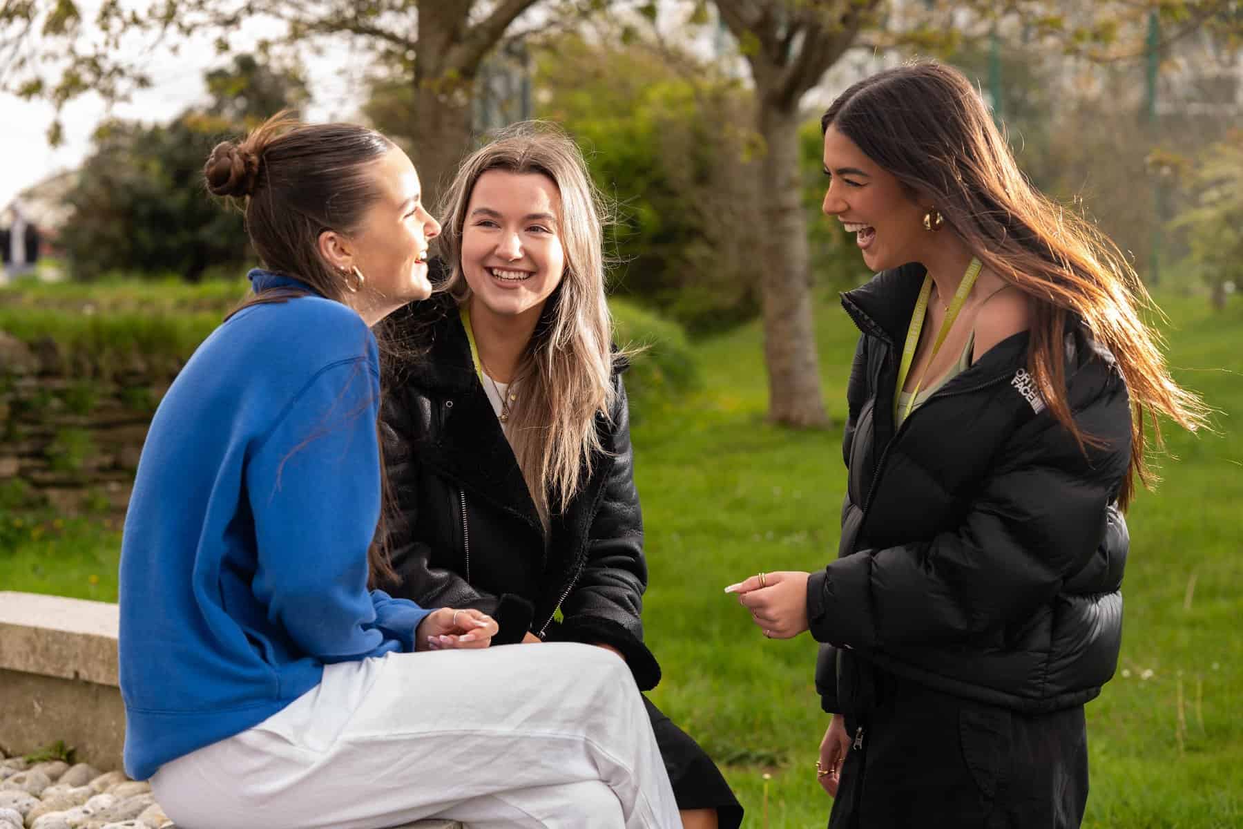General FE TC 057 Three girls are in a group outside, two sitting and one standing. They all laugh and smile.