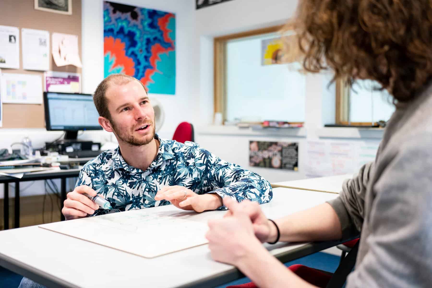 Humanities Social Sciences PC 006 A tutor kneels in front of a student who is sat at a classroom desk. The tutor is explaining something to the student and has a pen in their hand.