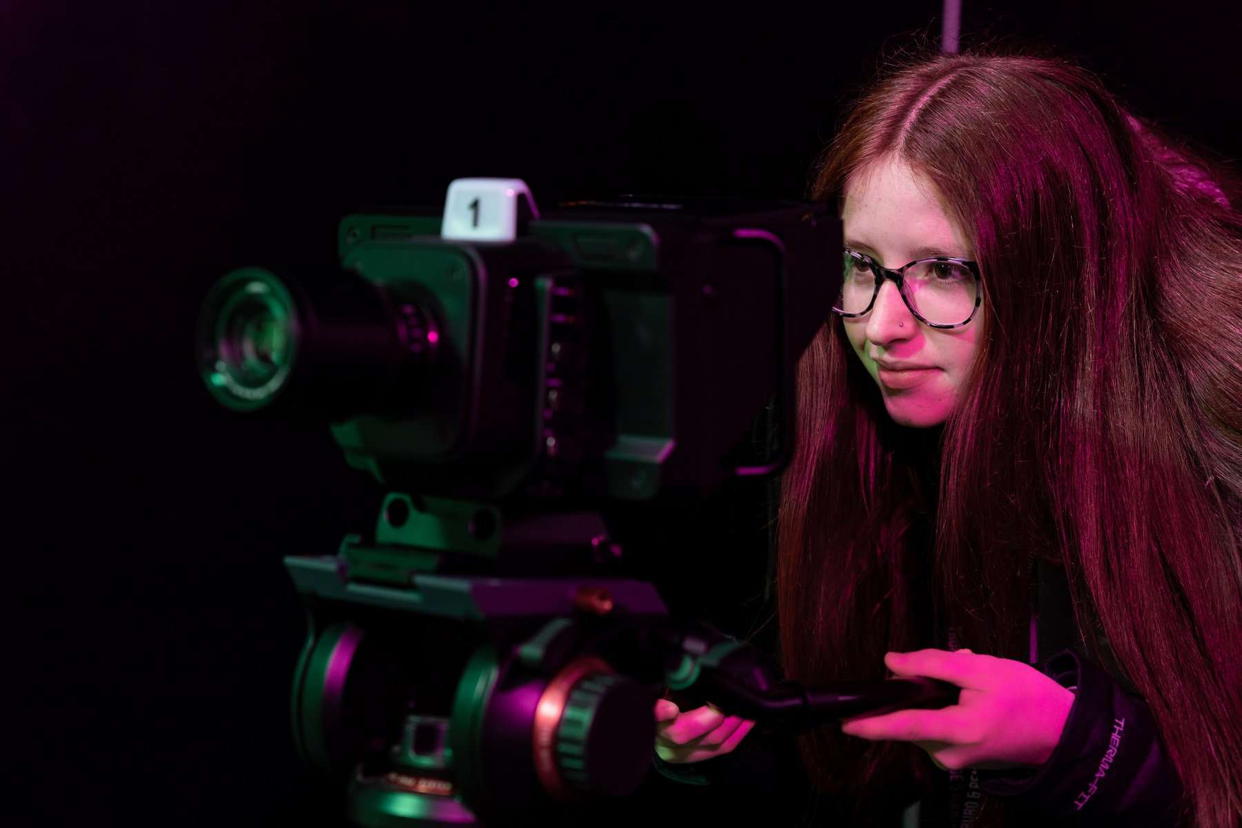 Media Photography HE 010 A student looks intently through a large digital camera. They have long red hair and glasses.